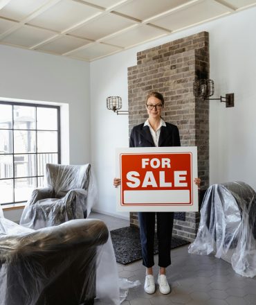Professional real estate agent holding a 'For Sale' sign in a furnished indoor setting.