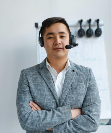 Asian call center agent wearing a headset and gray suit standing confidently indoors.